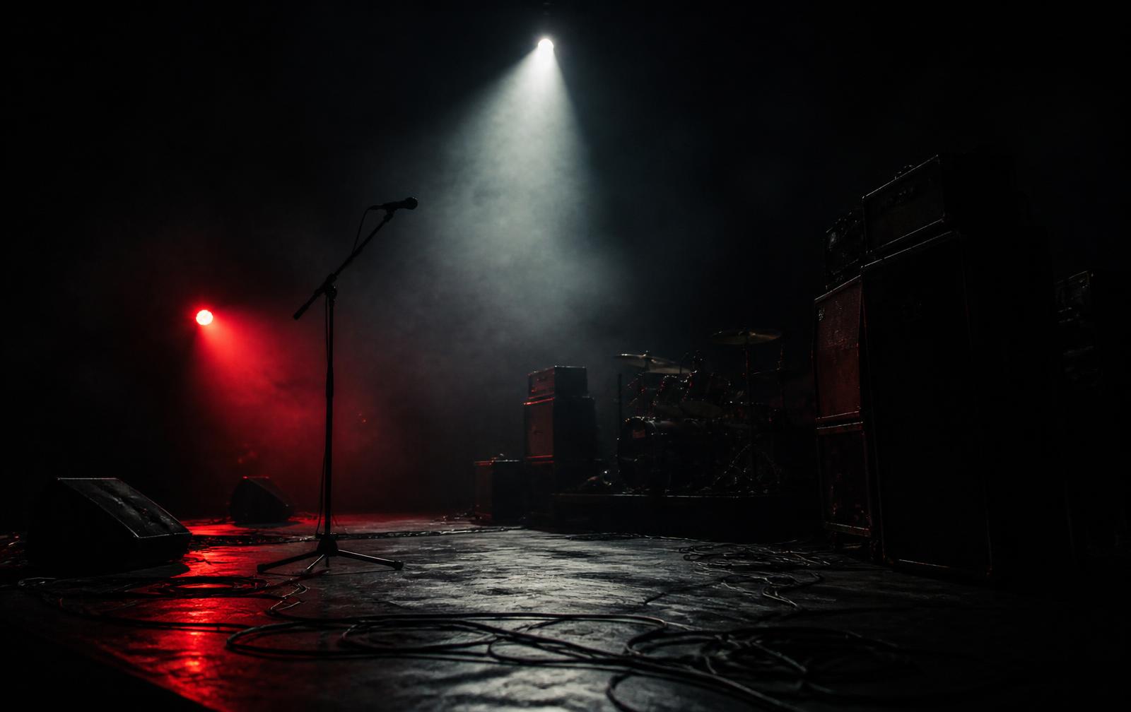 An empty, smoke-filled stage: amplifier stacks, a drum kit in shadow and a microphone with no one behind it